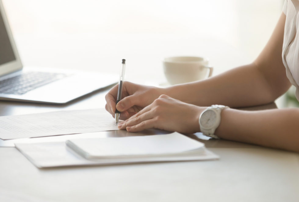 Close-up of a person’s hands filling out paperwork at a desk, with a pen, documents, a laptop, and a coffee cup in the background
