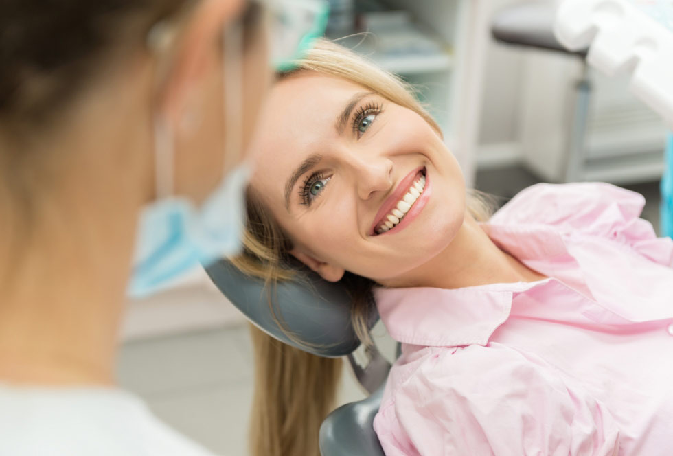 Smiling woman reclining in a dental chair, looking at a masked dental professional during a consultation or treatment in a modern clinic